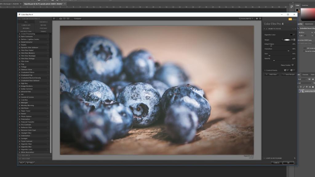 En computerskærm viser fotoredigeringssoftware med et nærbillede af blåbær på en træoverflade, der redigeres; redigeringsværktøjer, herunder muligheden for at tilføje vignet i Photoshop, og justeringspaneler er synlige rundt om billedet.